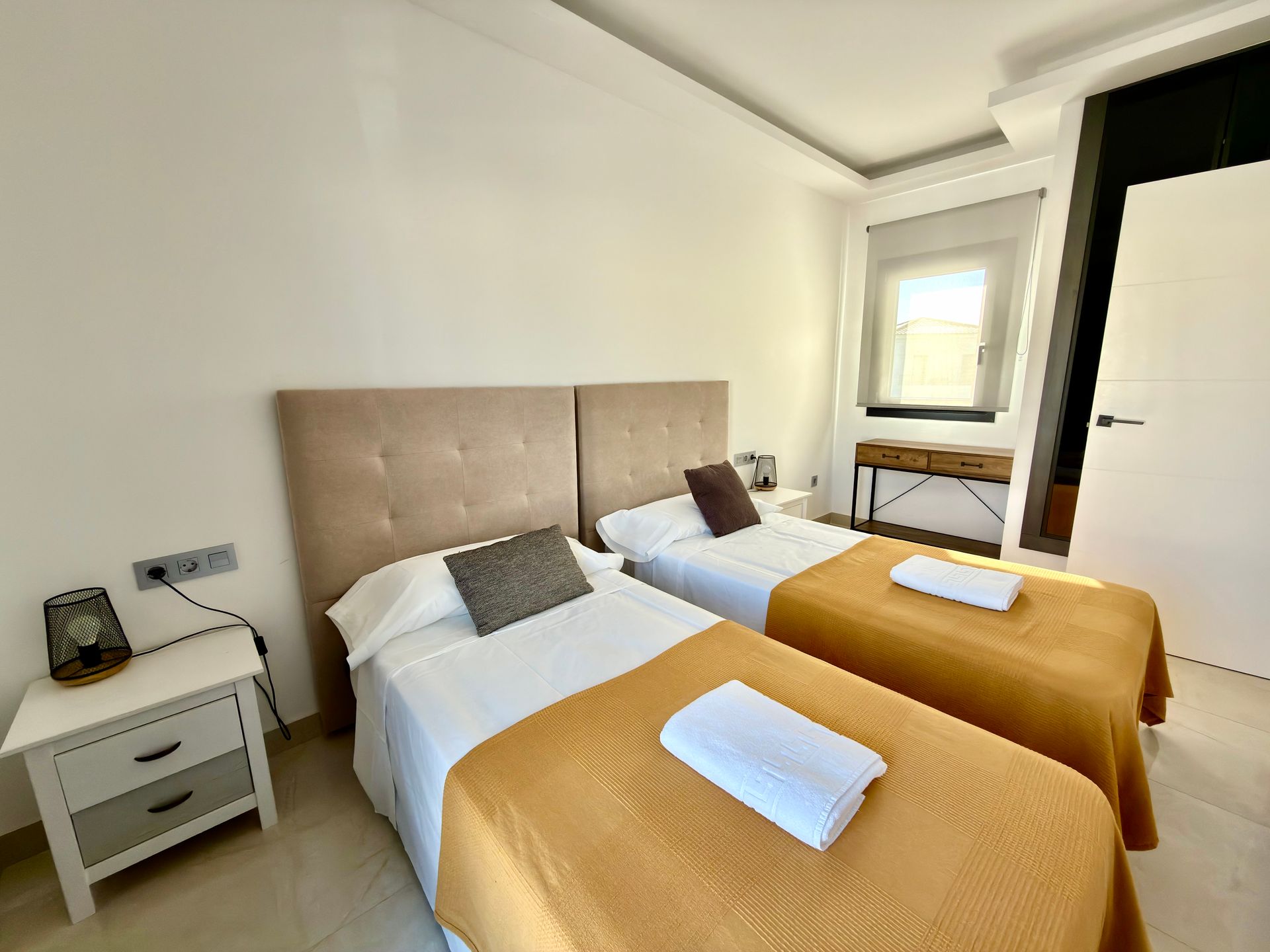 Habitación luminosa con dos camas individuales, mantas color mostaza, mesita de noche y ventana.