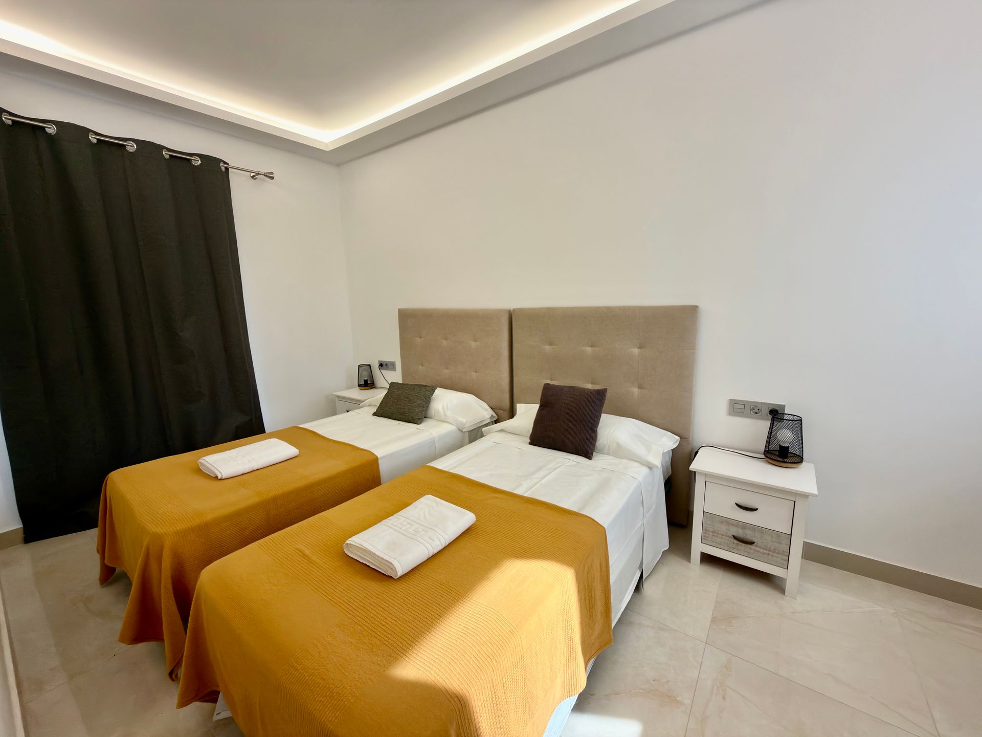 Habitación ordenada con dos camas individuales, colchas color mostaza, cortinas oscuras y una mesita de noche blanca.