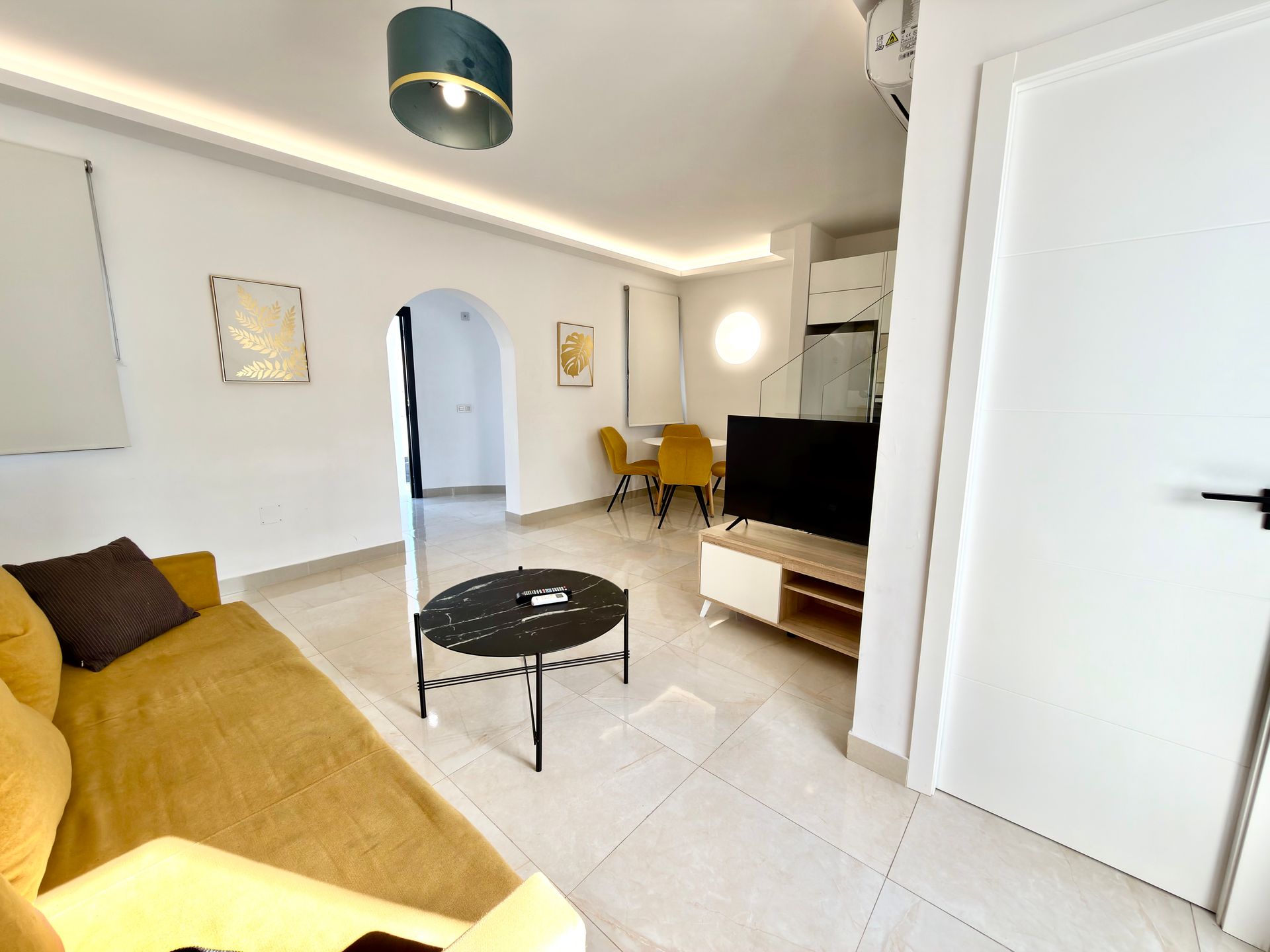 Sala de estar moderna con sofá color mostaza, mesa de centro negra, televisor, silla amarilla y puerta arqueada.