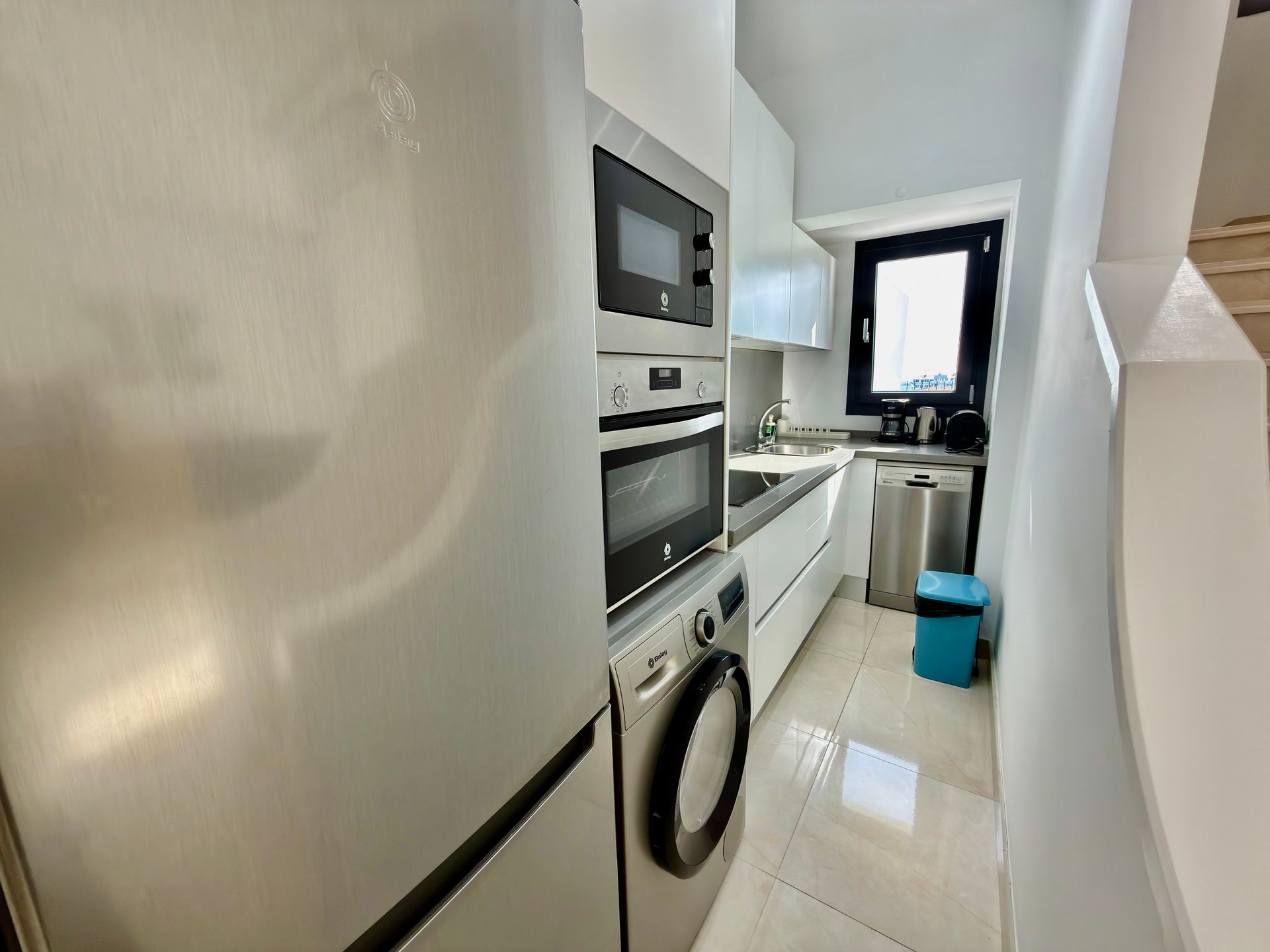Cocina estrecha de color blanco con horno empotrado, lavadora, fregadero y una ventana sobre la encimera.