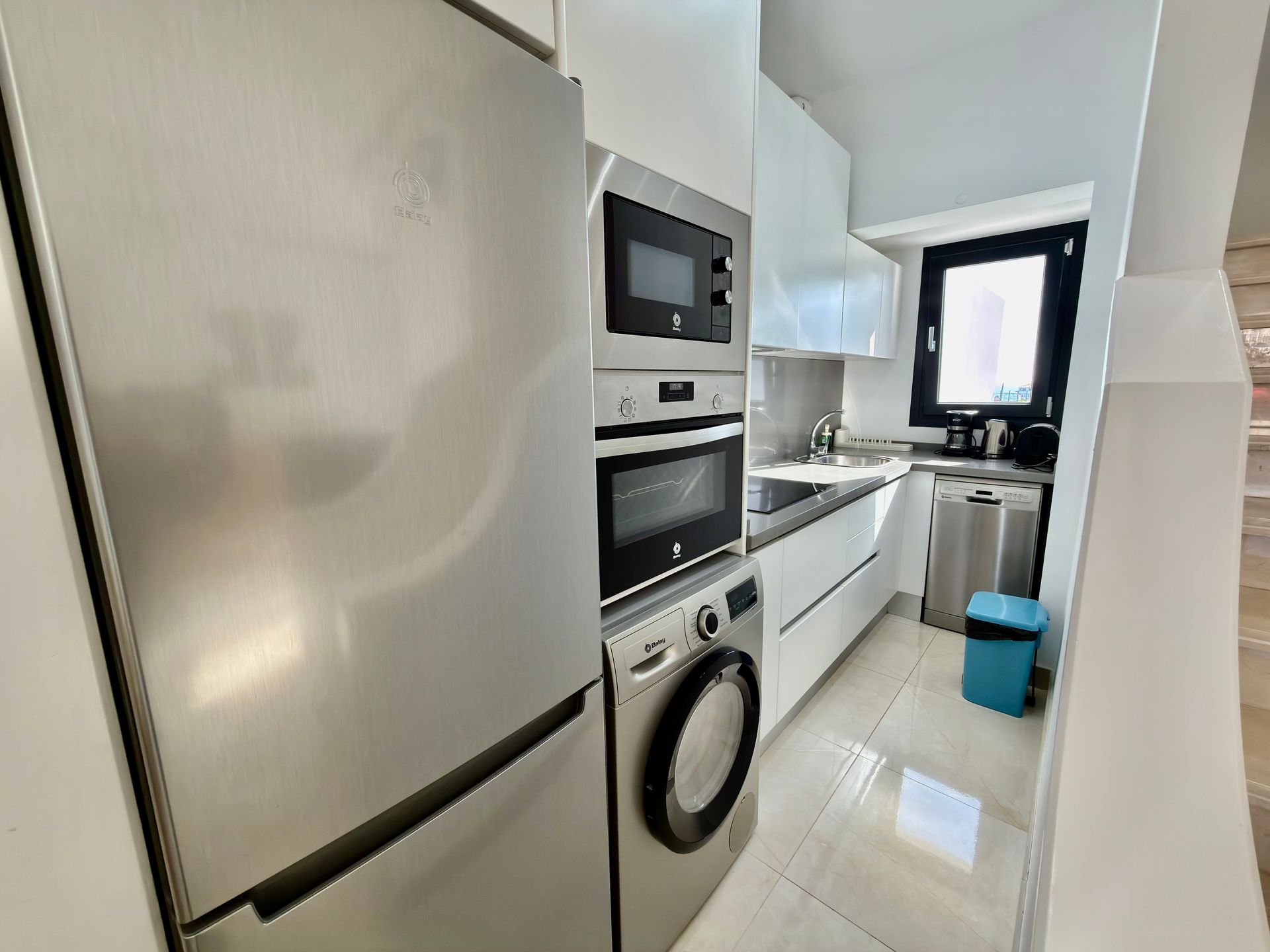 Cocina estrecha de color blanco con refrigerador, horno, lavadora, fregadero y ventana al final.