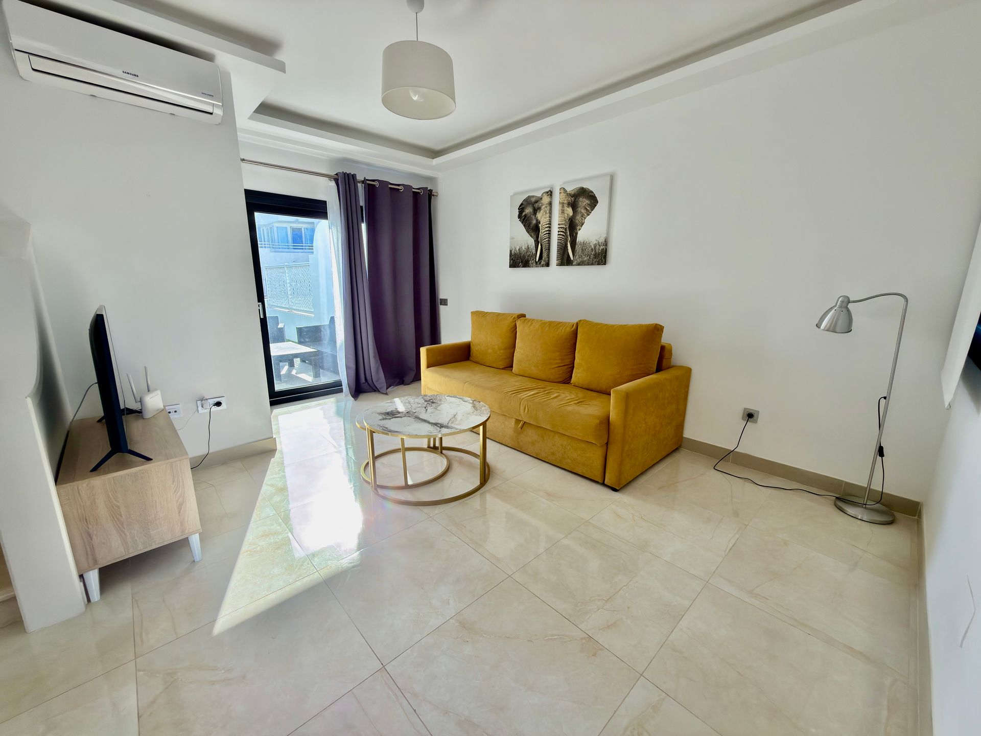 Sala de estar luminosa con sofá color mostaza, mesa de centro de cristal, televisor y puerta corredera con cortinas moradas.