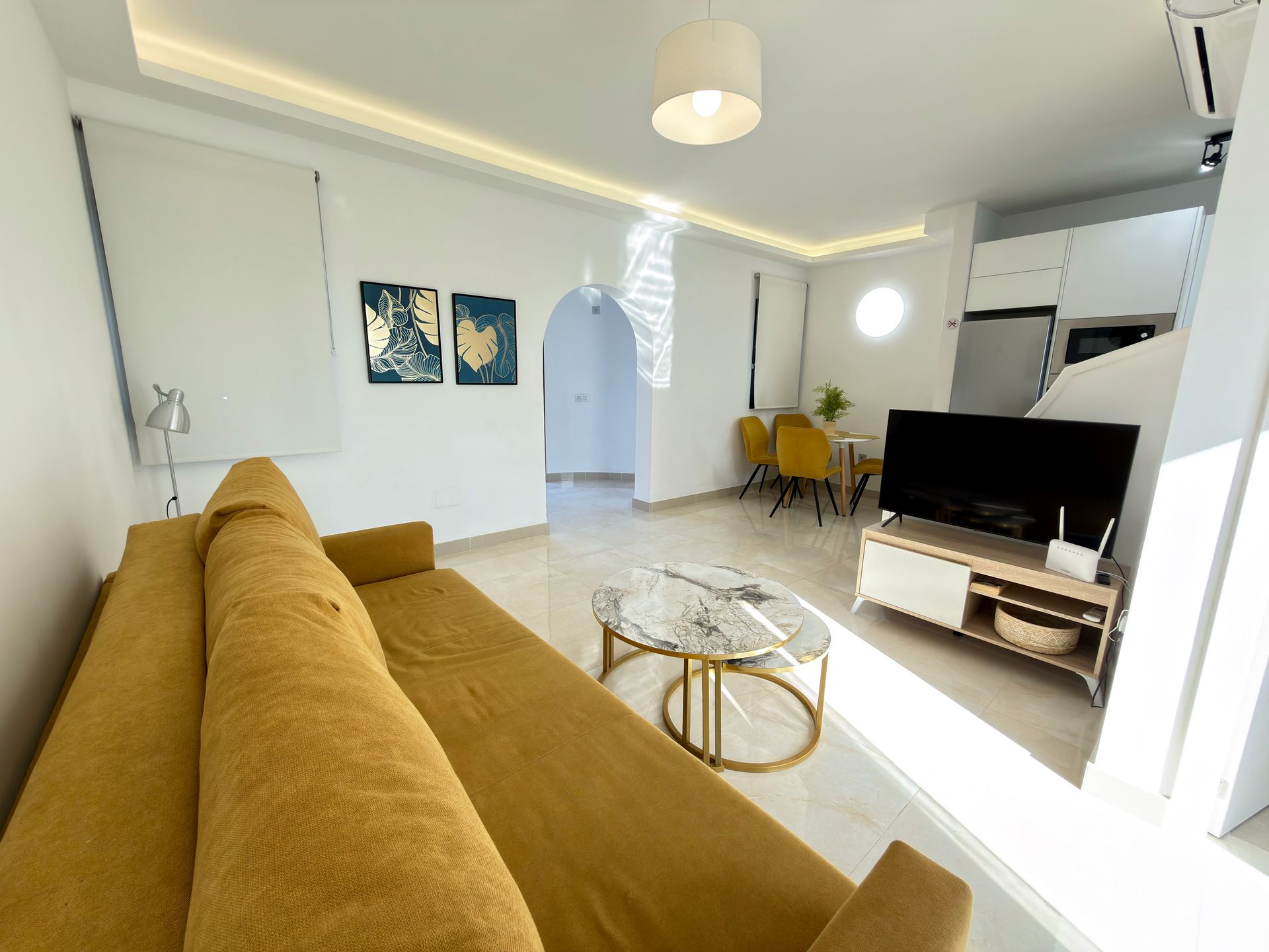 Sala de estar moderna blanca con sofá color mostaza, mesas de centro de cristal, televisor y zona de comedor.