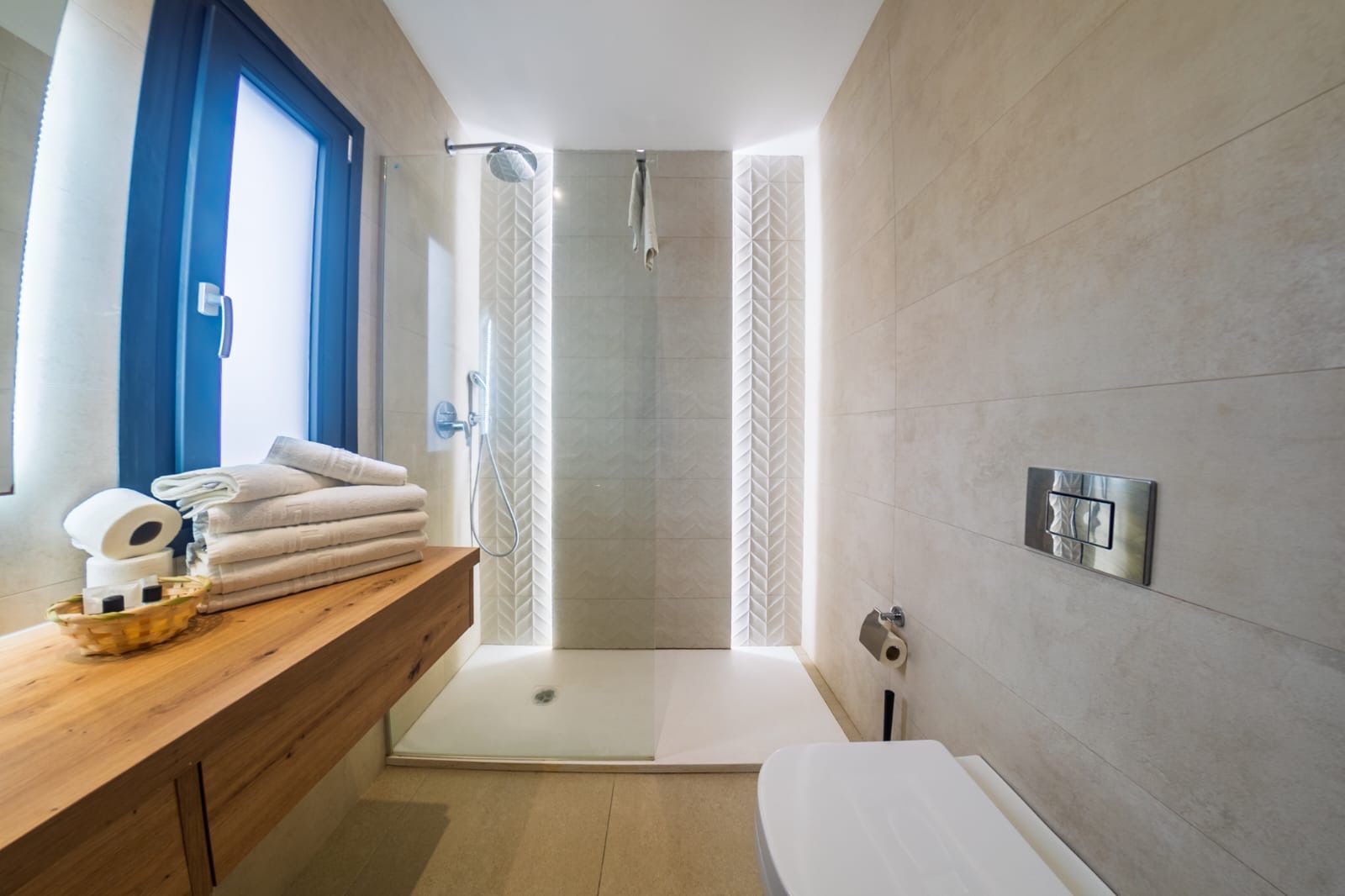 Baño moderno con ducha a ras de suelo, bañera, banco de madera y luz azul en la ventana.