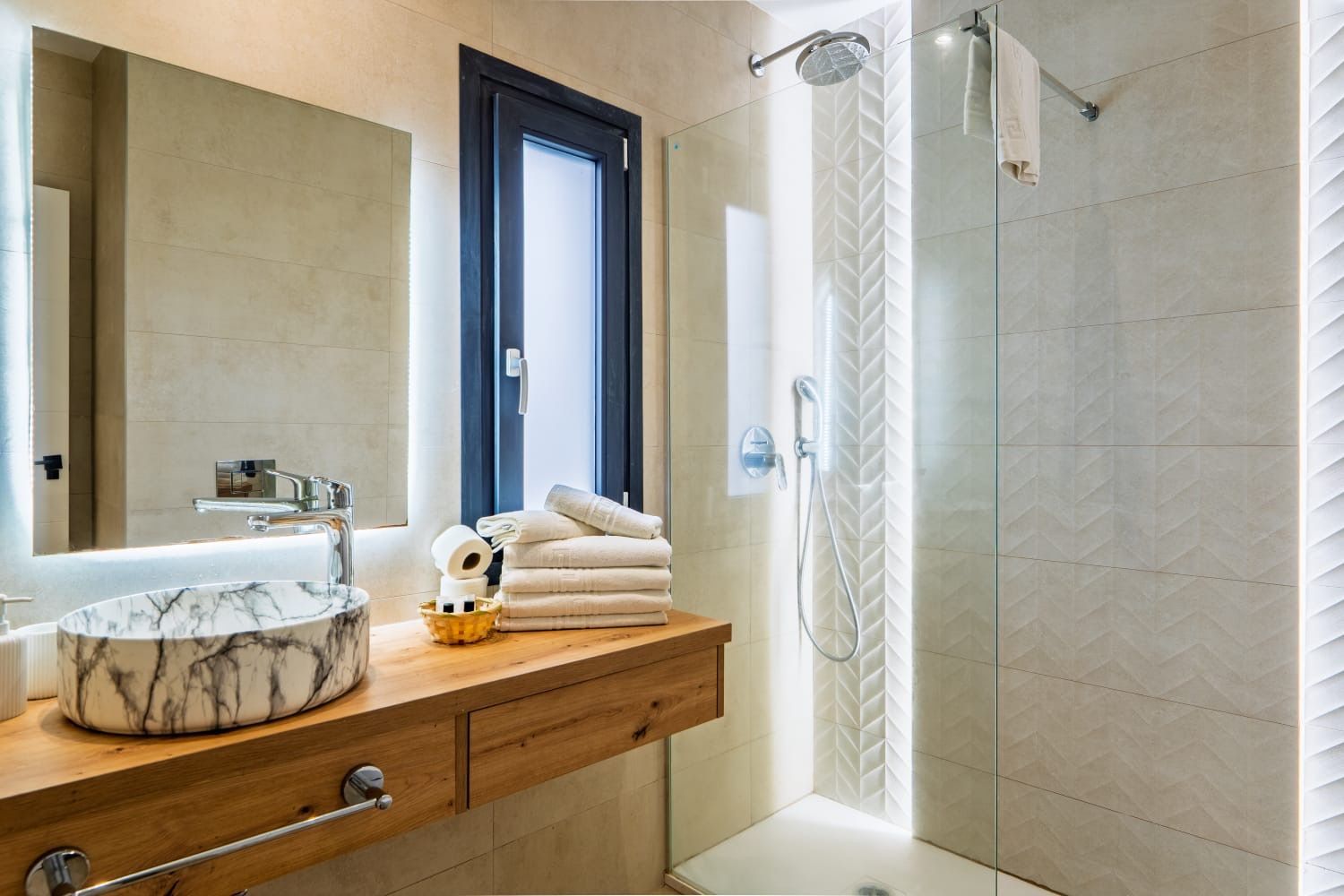 Baño moderno con tocador de madera, lavabo de mármol, espejo, toallas y mampara de ducha de cristal.
