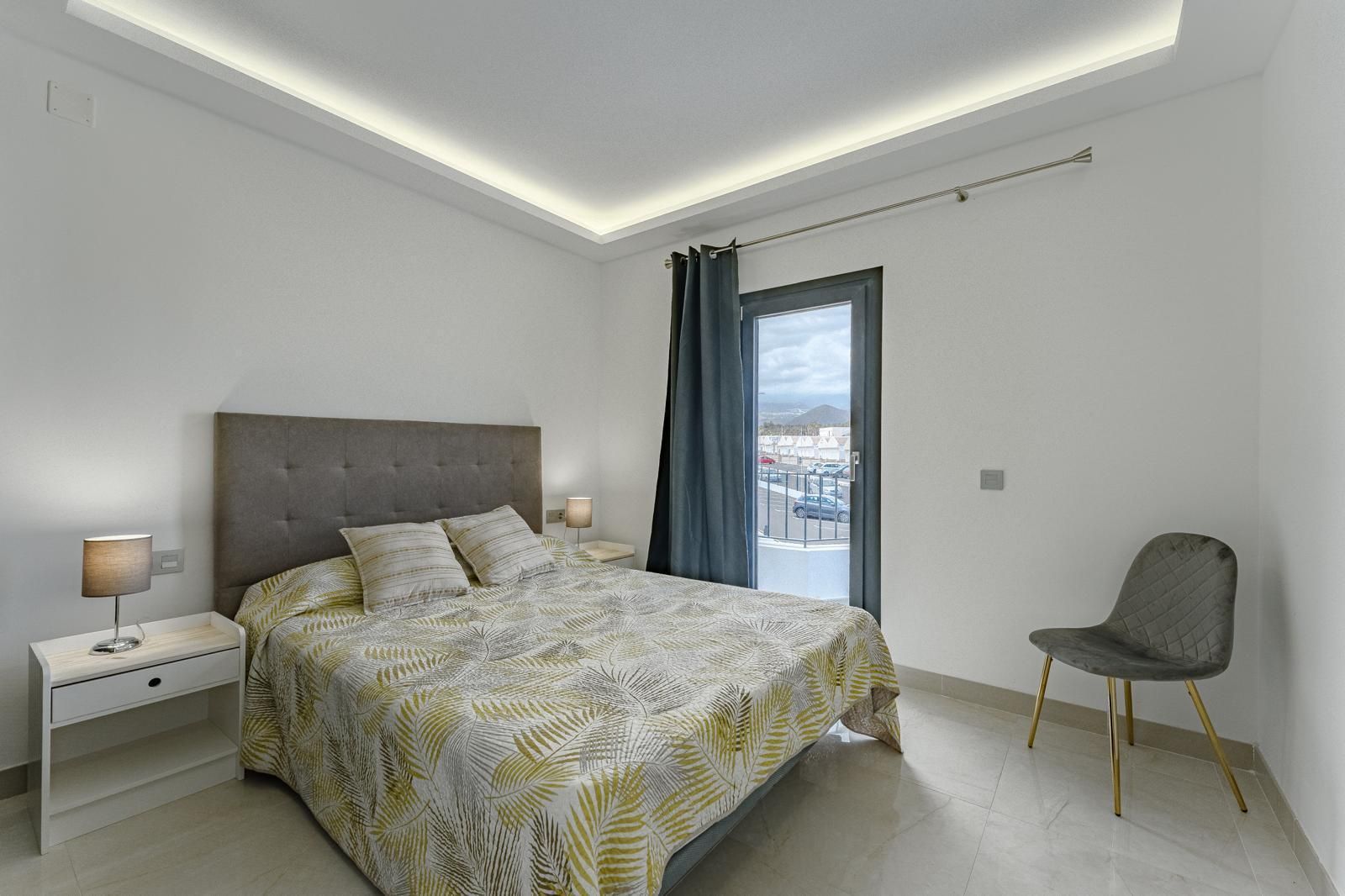 Habitación luminosa con cama, mesita de noche, silla y puerta corredera de cristal que da a un balcón.