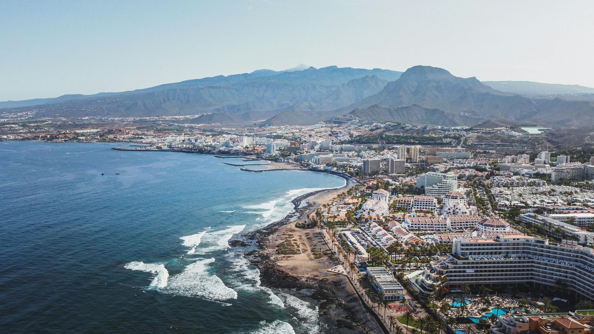 Vista aérea de una ciudad costera con una playa curva, océano azul y montañas en la distancia.