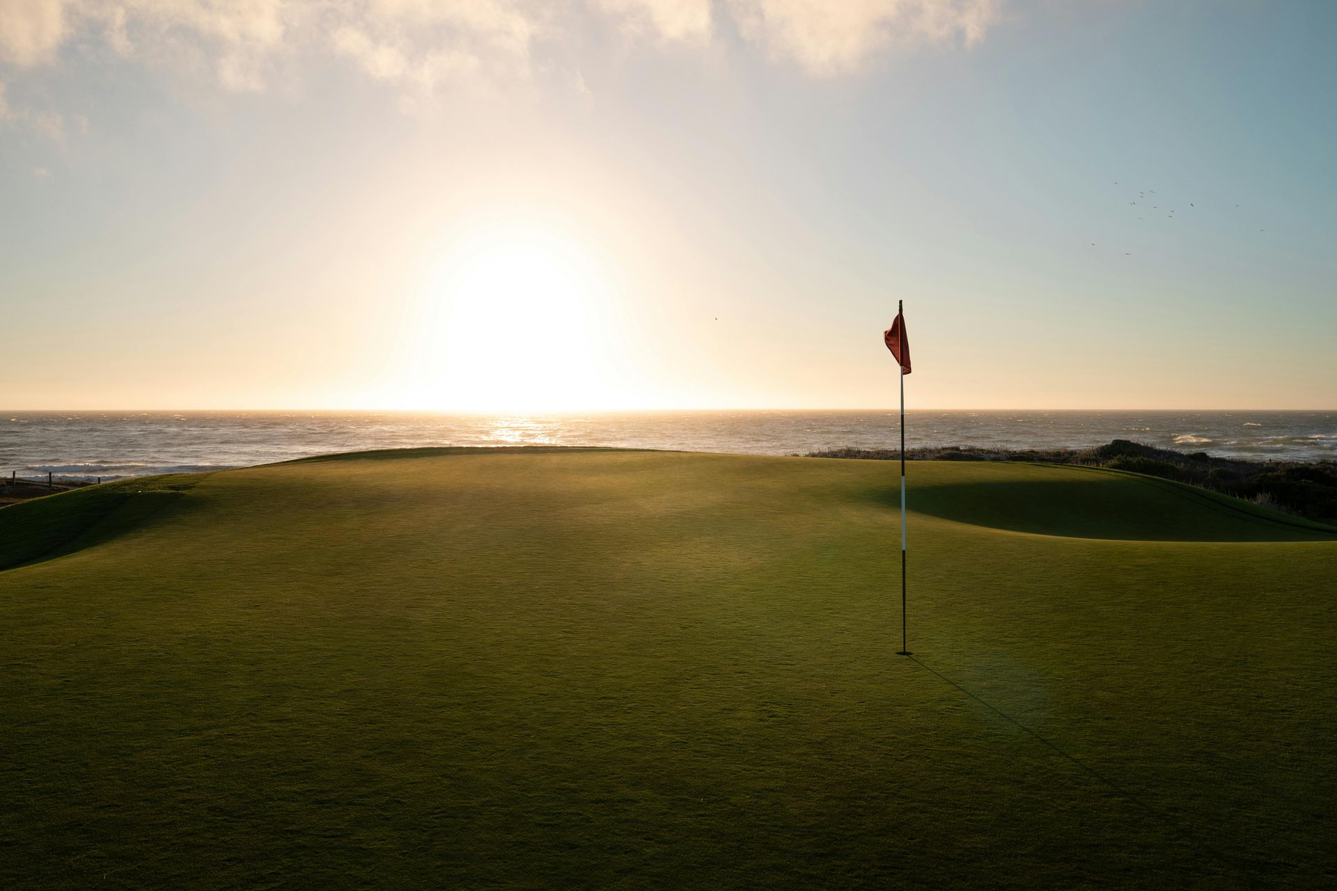 Campo de golf iluminado por el sol junto al océano con una bandera al atardecer.