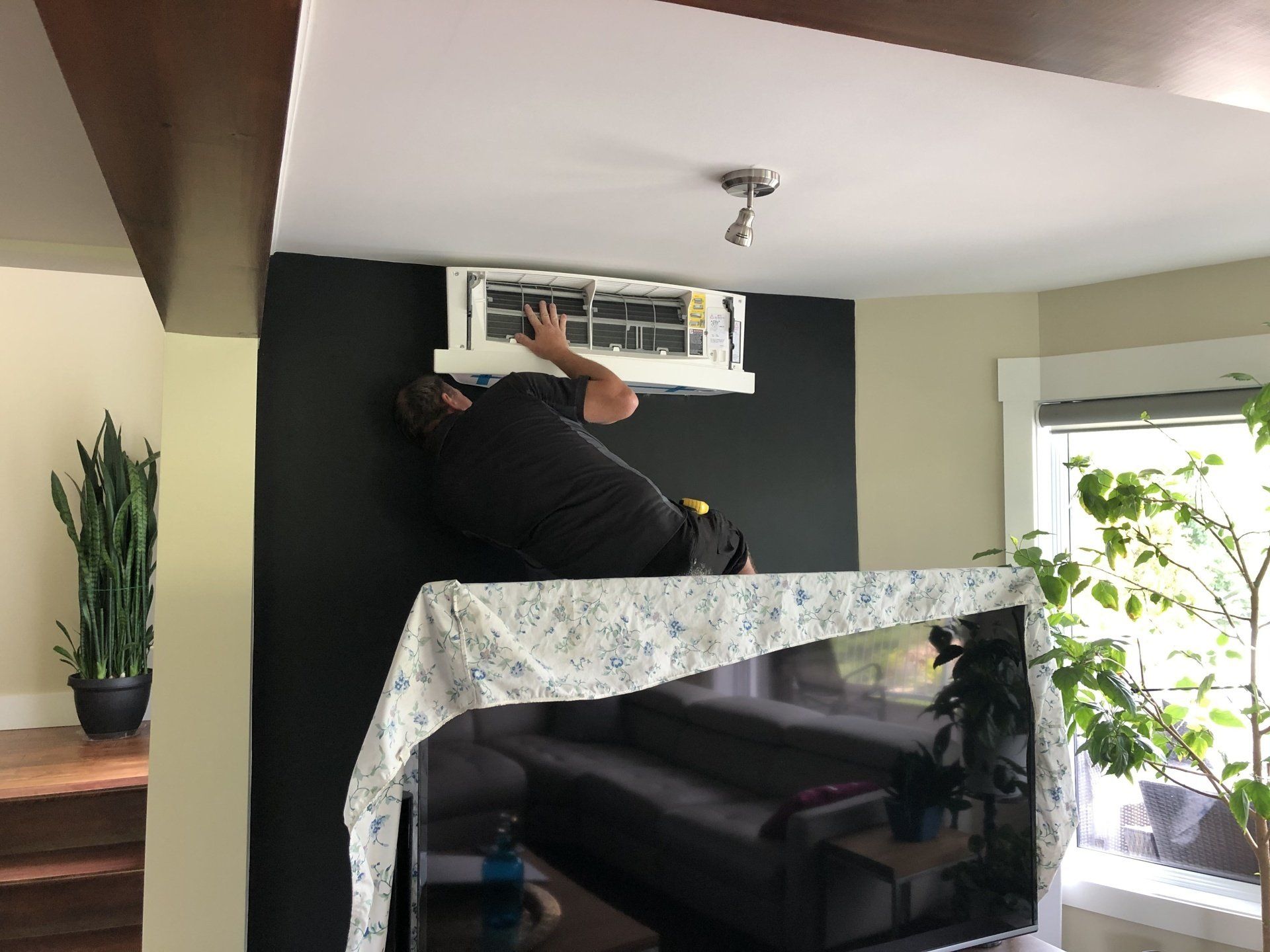 Un homme travaille sur un climatiseur fixé à un mur noir, au-dessus d'un téléviseur recouvert.