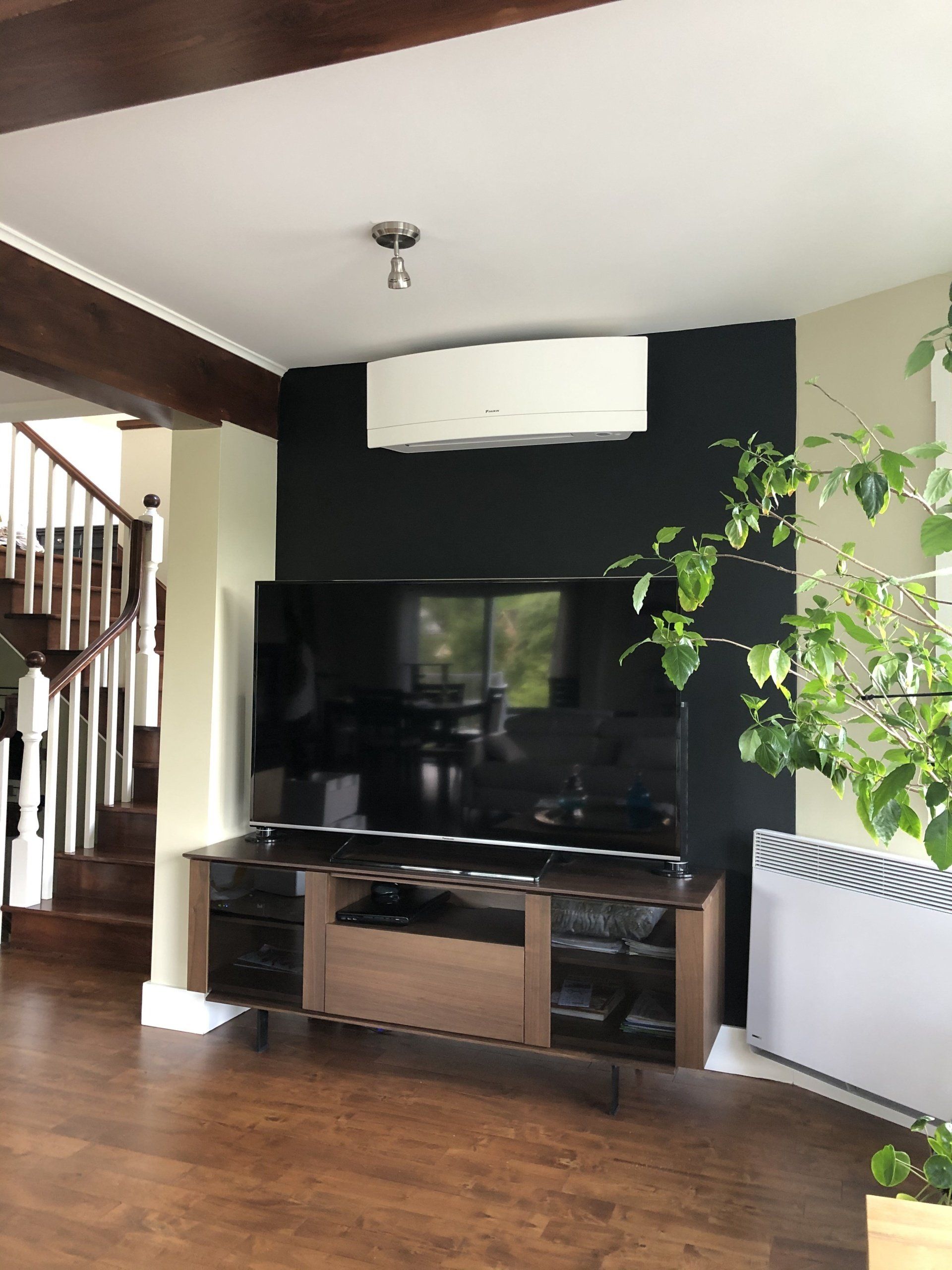 Salon avec télévision sur un mur sombre, climatiseur au-dessus et escalier.