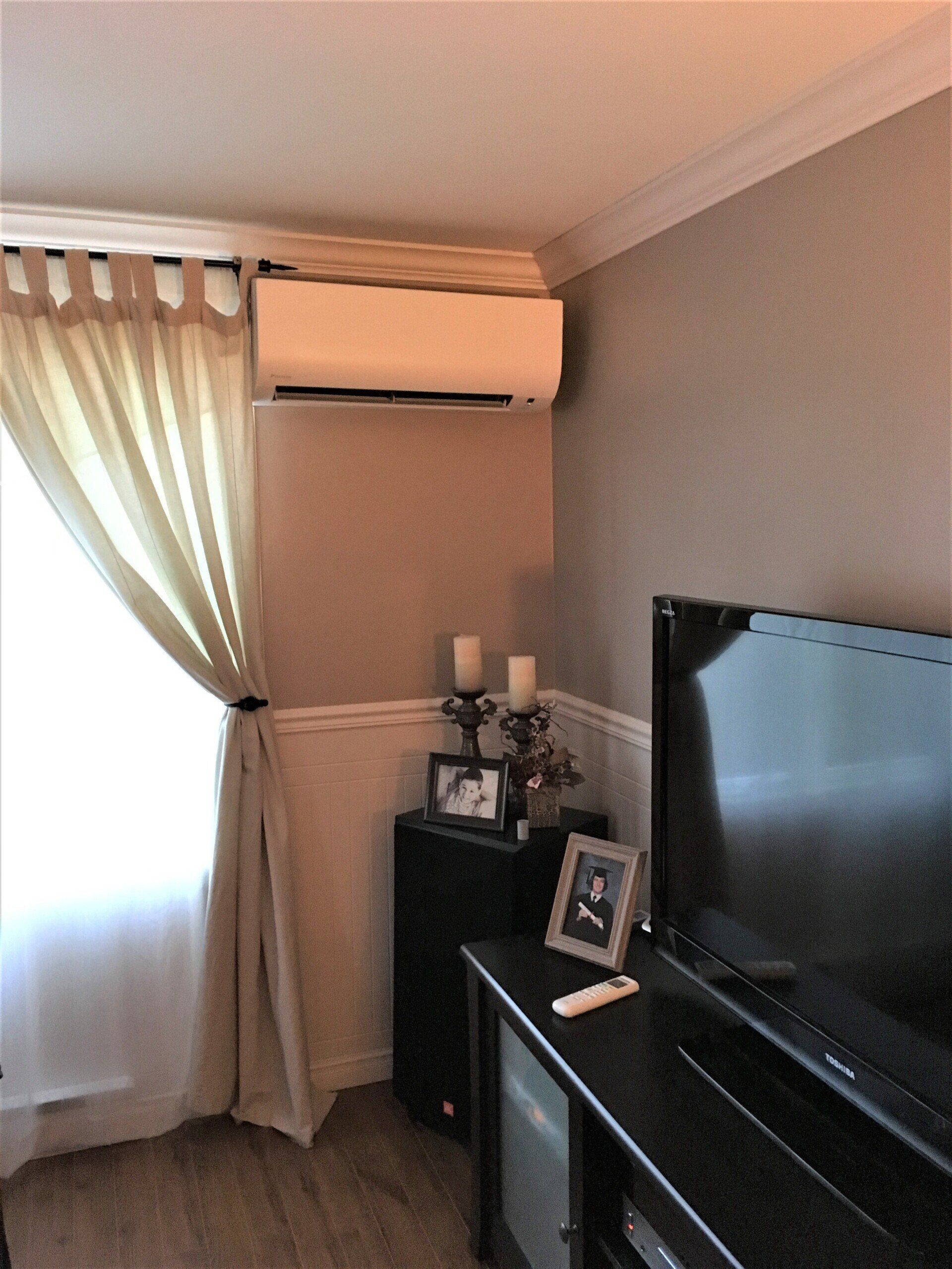 Climatiseur fixé sur un mur gris au-dessus d'un meuble noir avec une télévision et une fenêtre aux rideaux beiges.