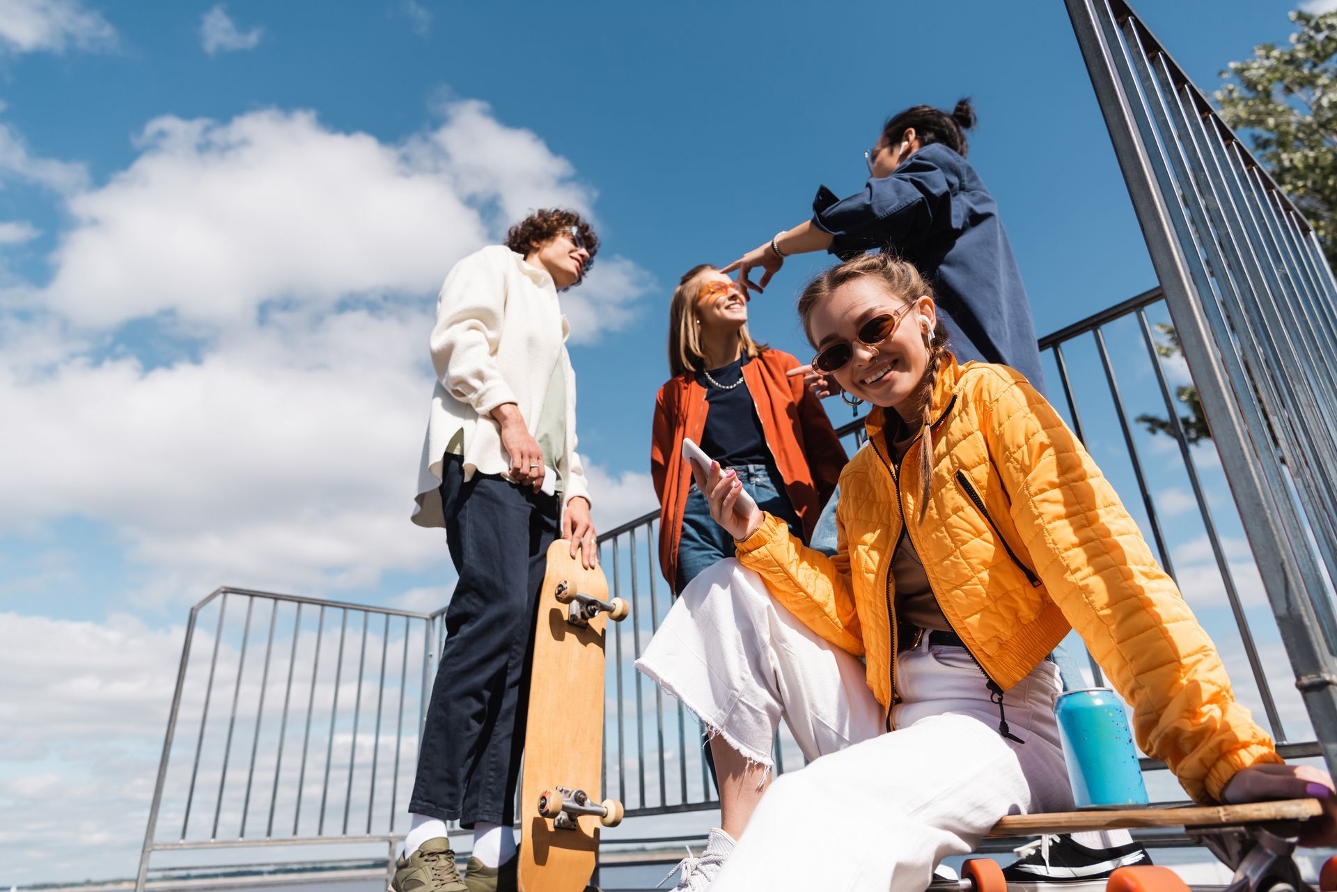 Group of young people with skateboards, posing outdoors. One person is sitting, others are standing; blue sky background.
