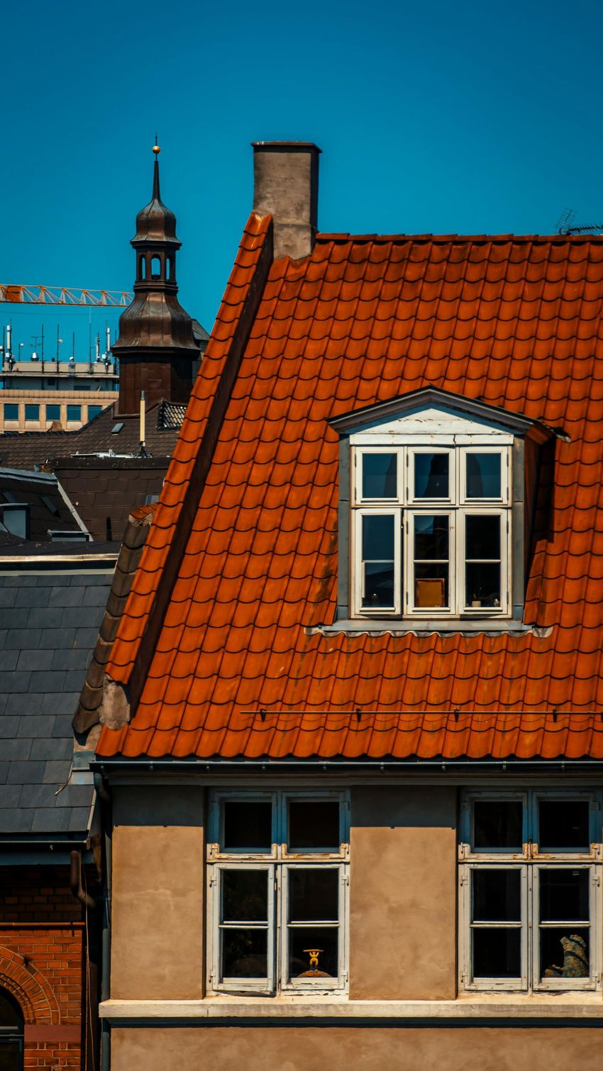 Red-tiled rooftop with dormer windows against a blue sky, with a church tower in the background
