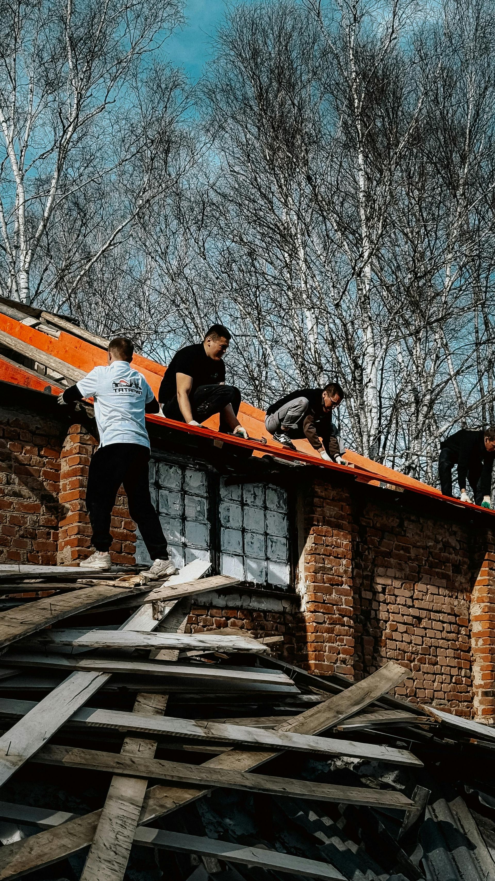 People working on a damaged roof beside a brick wall and collapsed wooden debris under bare trees