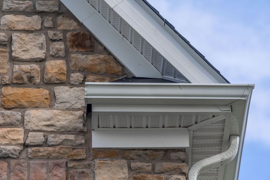 Stone house roof eave with white gutter and downspout against a blue sky