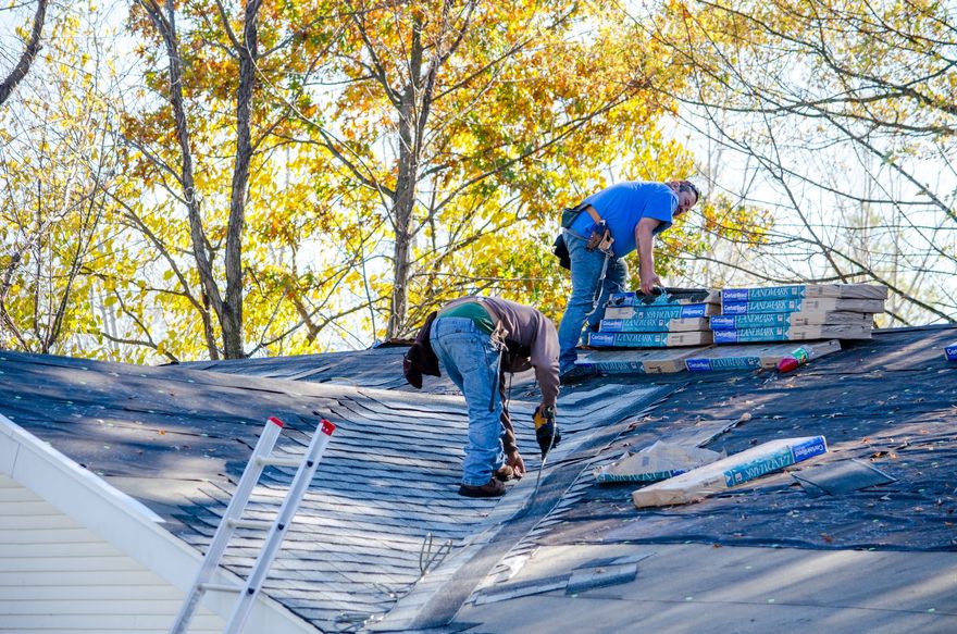 Workers repairing a blue roof with ladders amid autumn trees