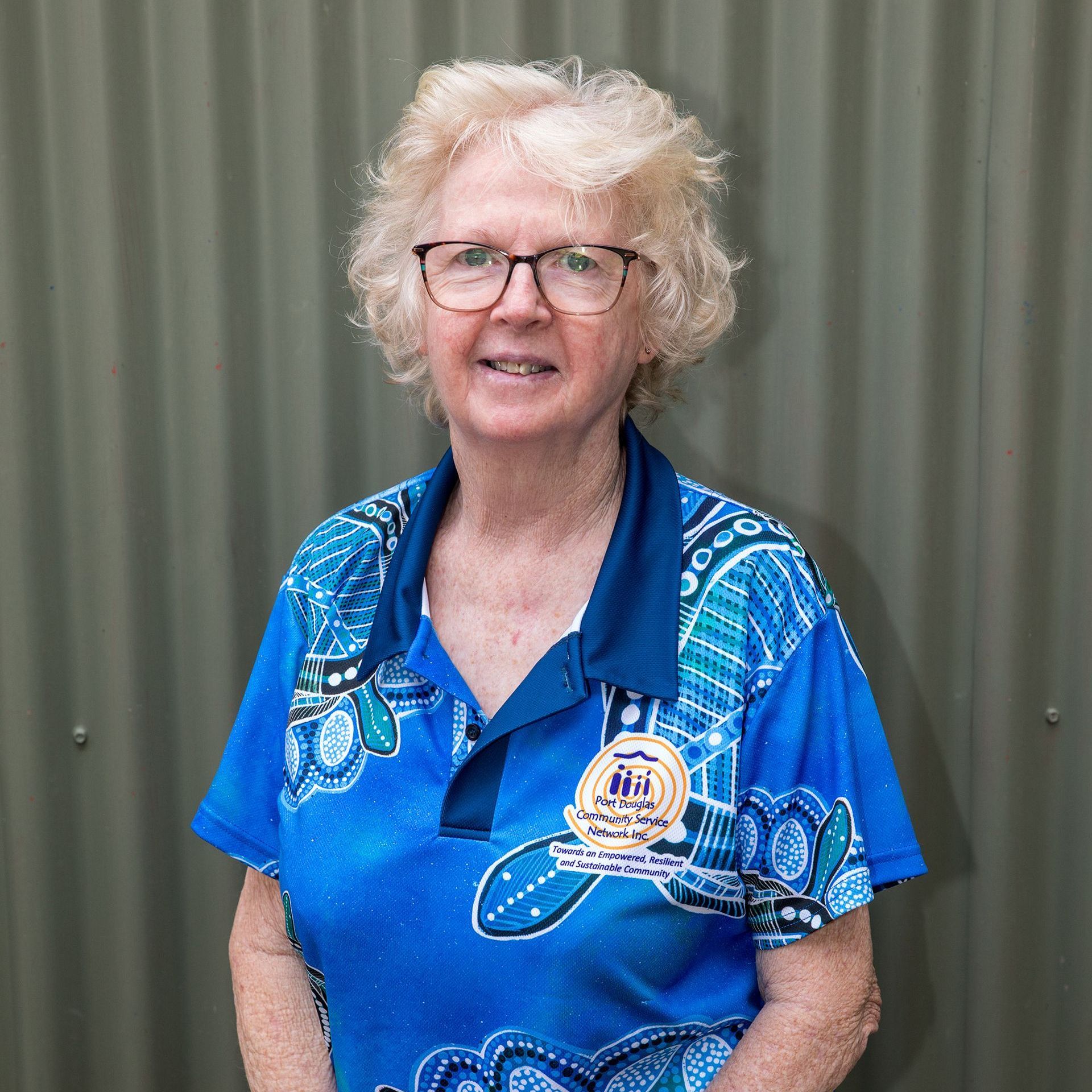 A woman wearing glasses and a blue shirt is standing in front of a green wall.