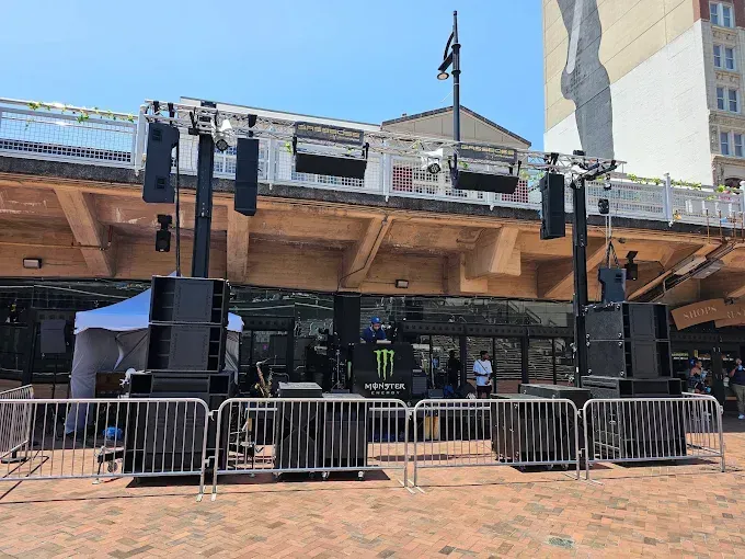 Outdoor stage setup with sound equipment and lights, metal barriers in front of a building with people.
