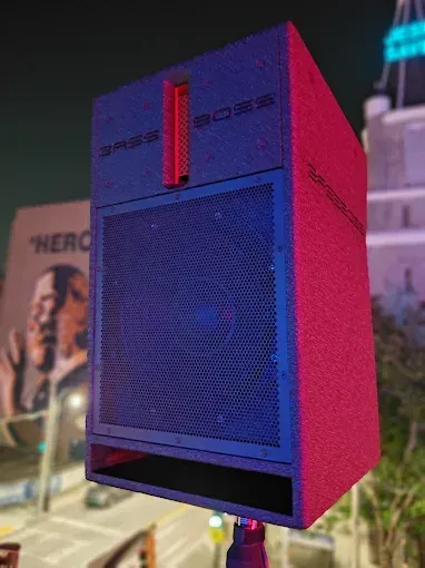 A black speaker box with a mesh front, red accents, mounted outdoors with a mural in the background.