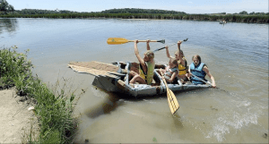 People in homemade boat, paddling on a lake, raising paddles. Blue and yellow life vests. Sunny day.