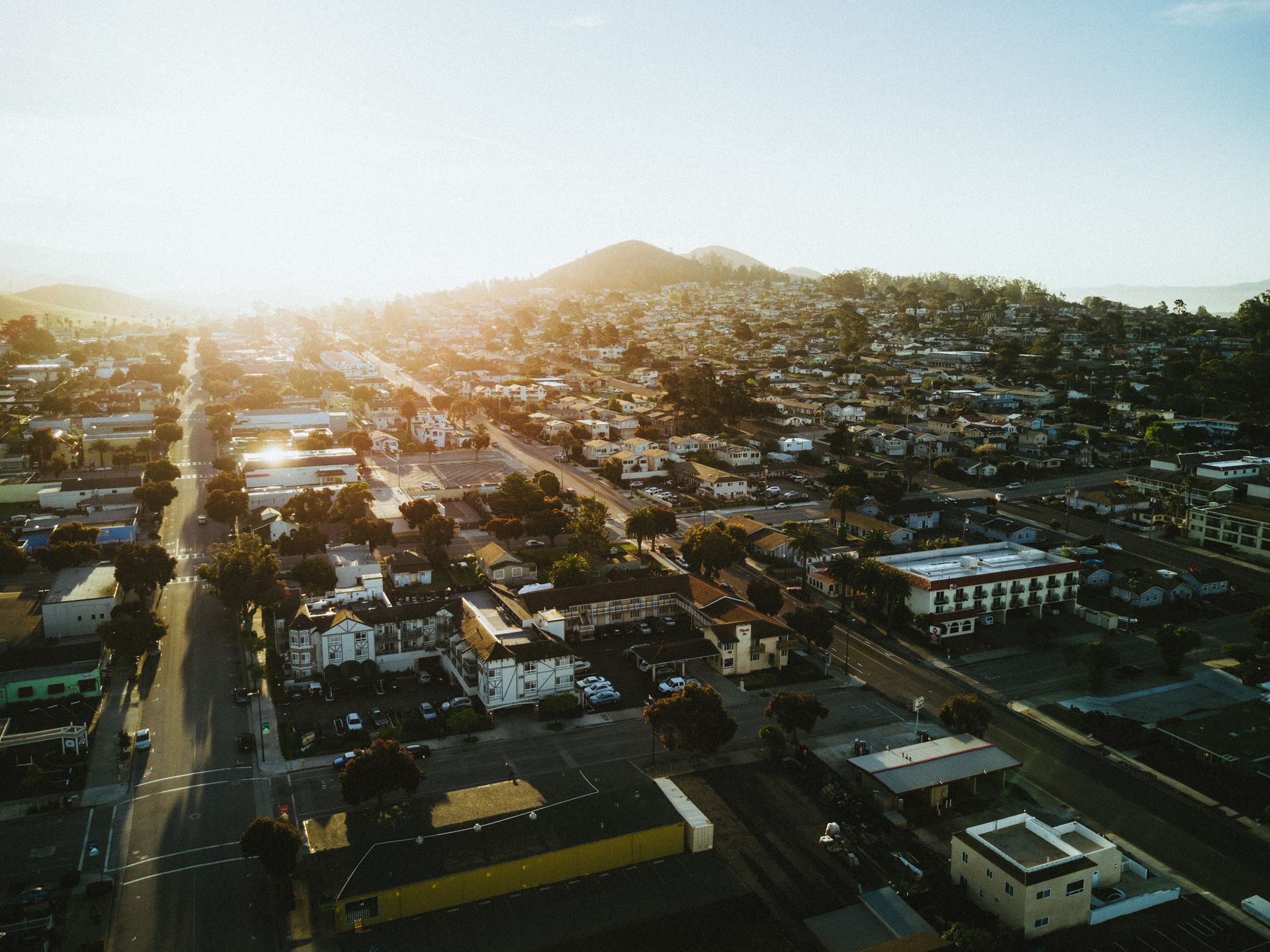 Aerial view of a sunlit suburban neighborhood with houses and buildings, hills in the background.