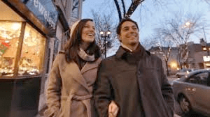 Couple strolling on city street, arm in arm, smiling. Shop window, cars, and streetlights visible.