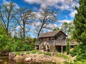 A rustic wooden watermill with a water wheel, set among green trees and a flowing river, under a blue sky.