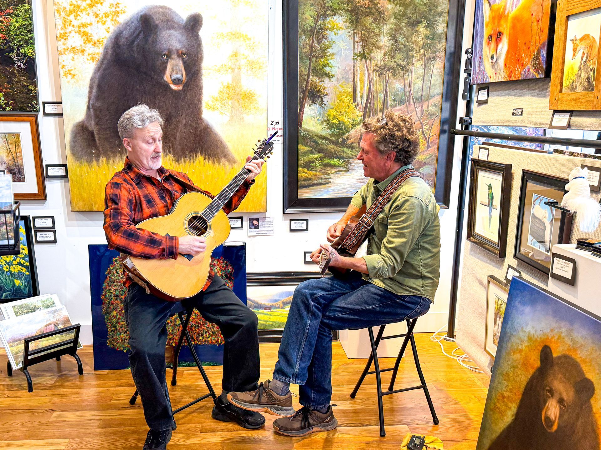 Two men sitting and playing acoustic guitars in an art gallery with bear paintings on the walls