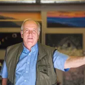 Man in a blue shirt and olive vest standing indoors with one arm outstretched