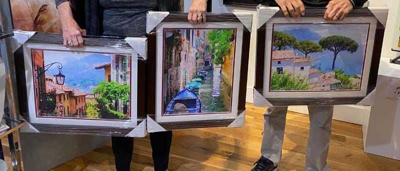 Three people holding framed paintings, including a street scene, a garden path, and a lakeside view.