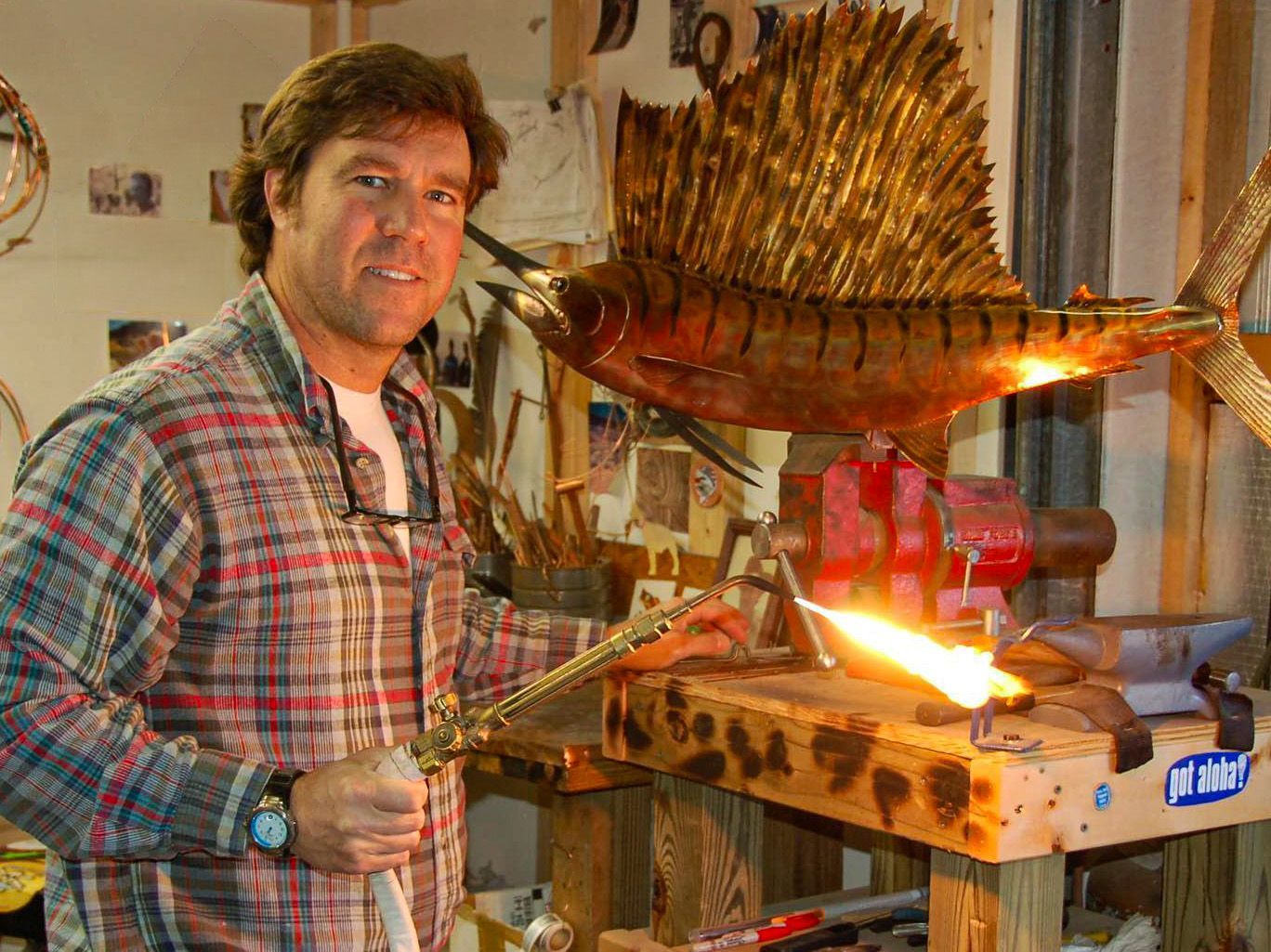 Man posing beside a metal fish sculpture with a small flame in a workshop