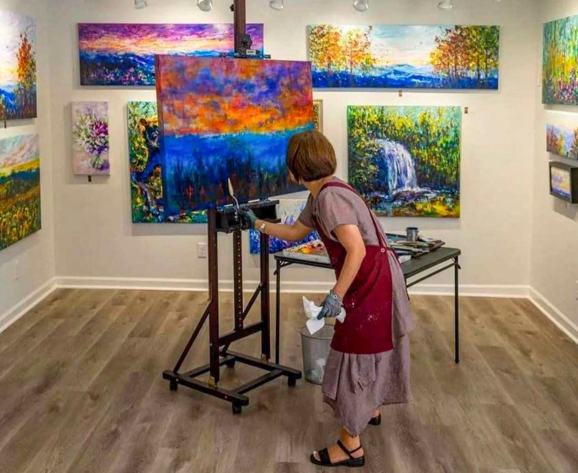 Artist painting colorful abstract canvas in a gallery with several bright landscape paintings on the walls