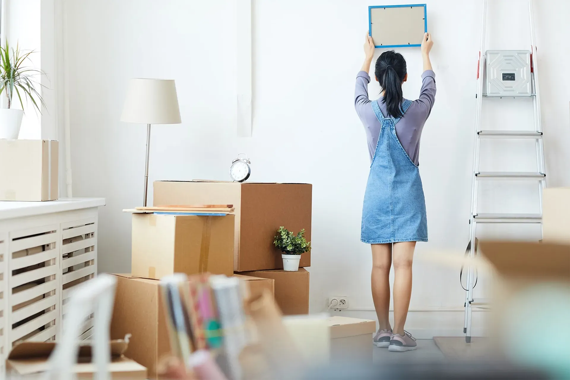 Woman in blue dress hangs a framed picture in a bright, moving boxes-filled room