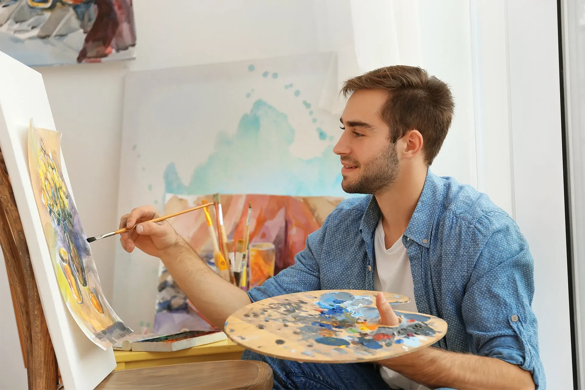 Artist painting on canvas, holding a palette in a bright studio.