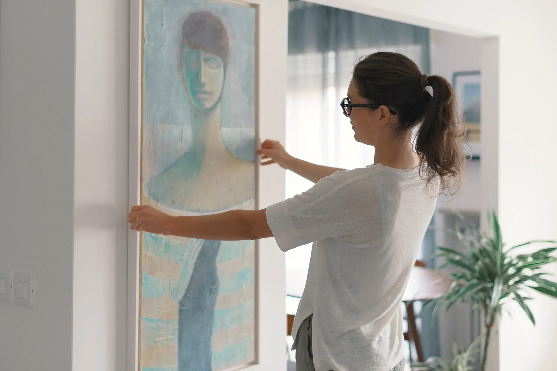 Woman hanging a framed abstract portrait on a wall in a bright room