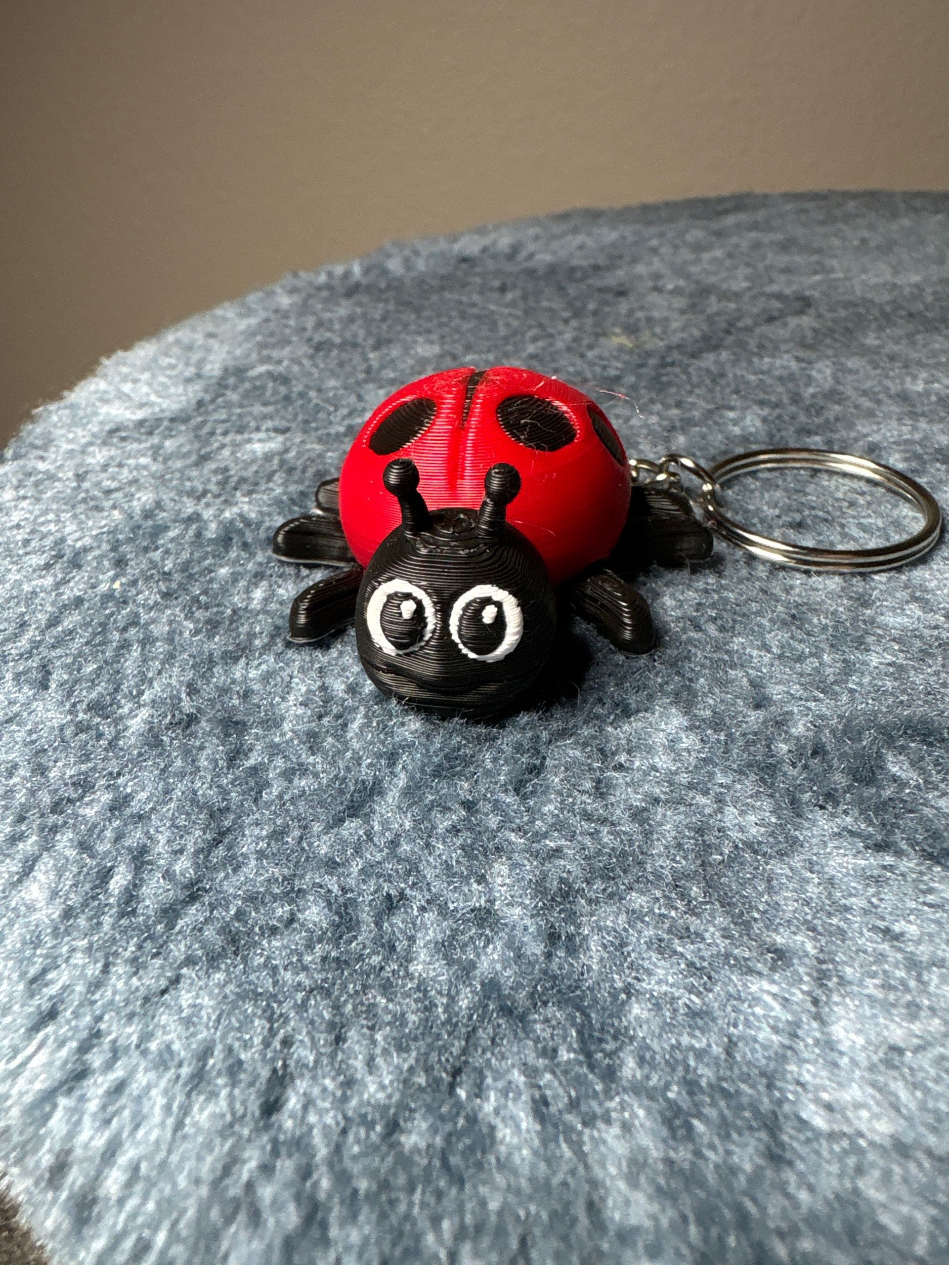A red and black 3D-printed ladybug keychain with white-painted eyes, sitting on a textured grey surface.