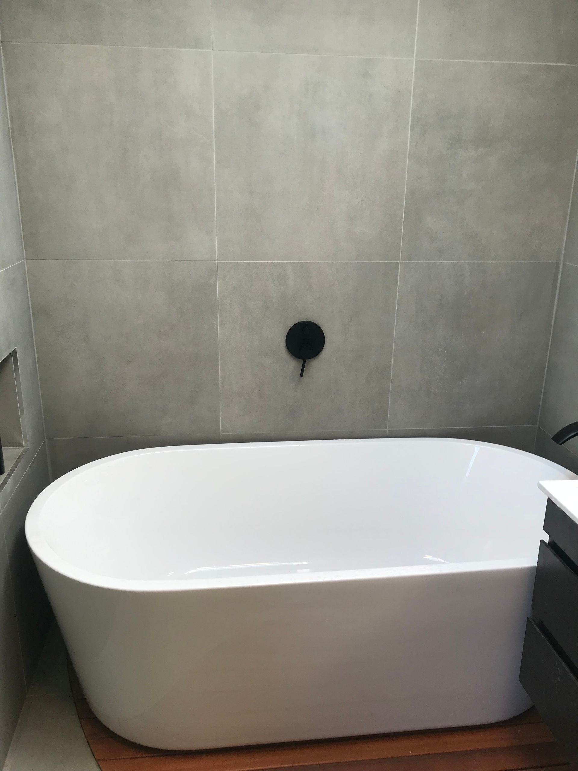 White freestanding bathtub against gray tiled bathroom wall with a black wall fixture