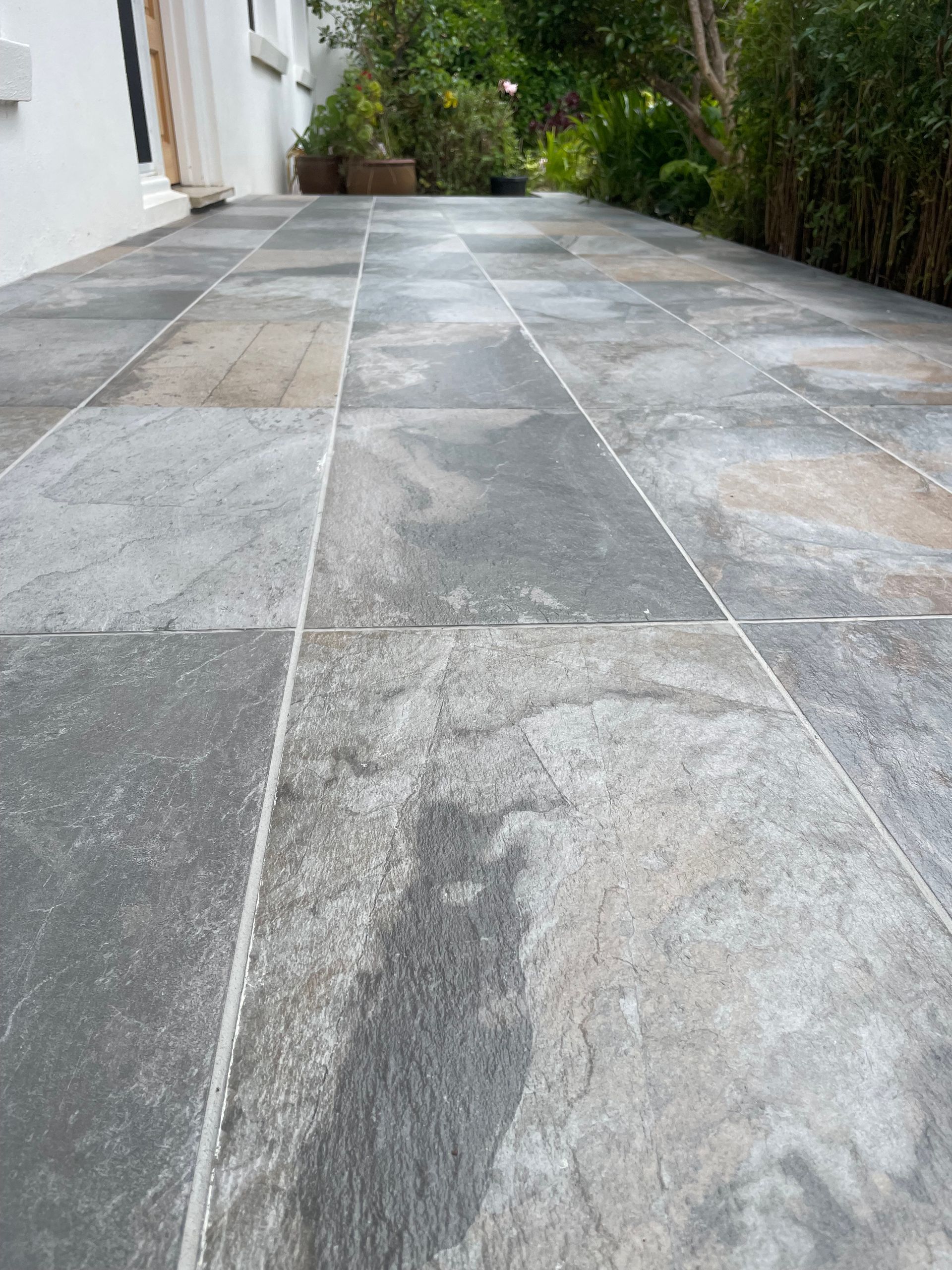 Gray stone patio walkway beside a white house, leading past green shrubs and trees Gray stone patio walkway beside a white house, leading past green shrubs and trees