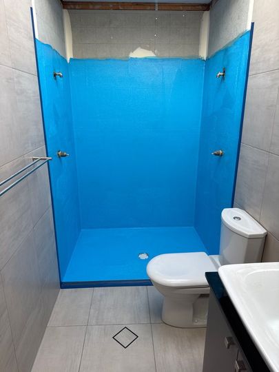 Small bathroom with a blue-tiled shower stall, toilet, and sink in the foreground.
