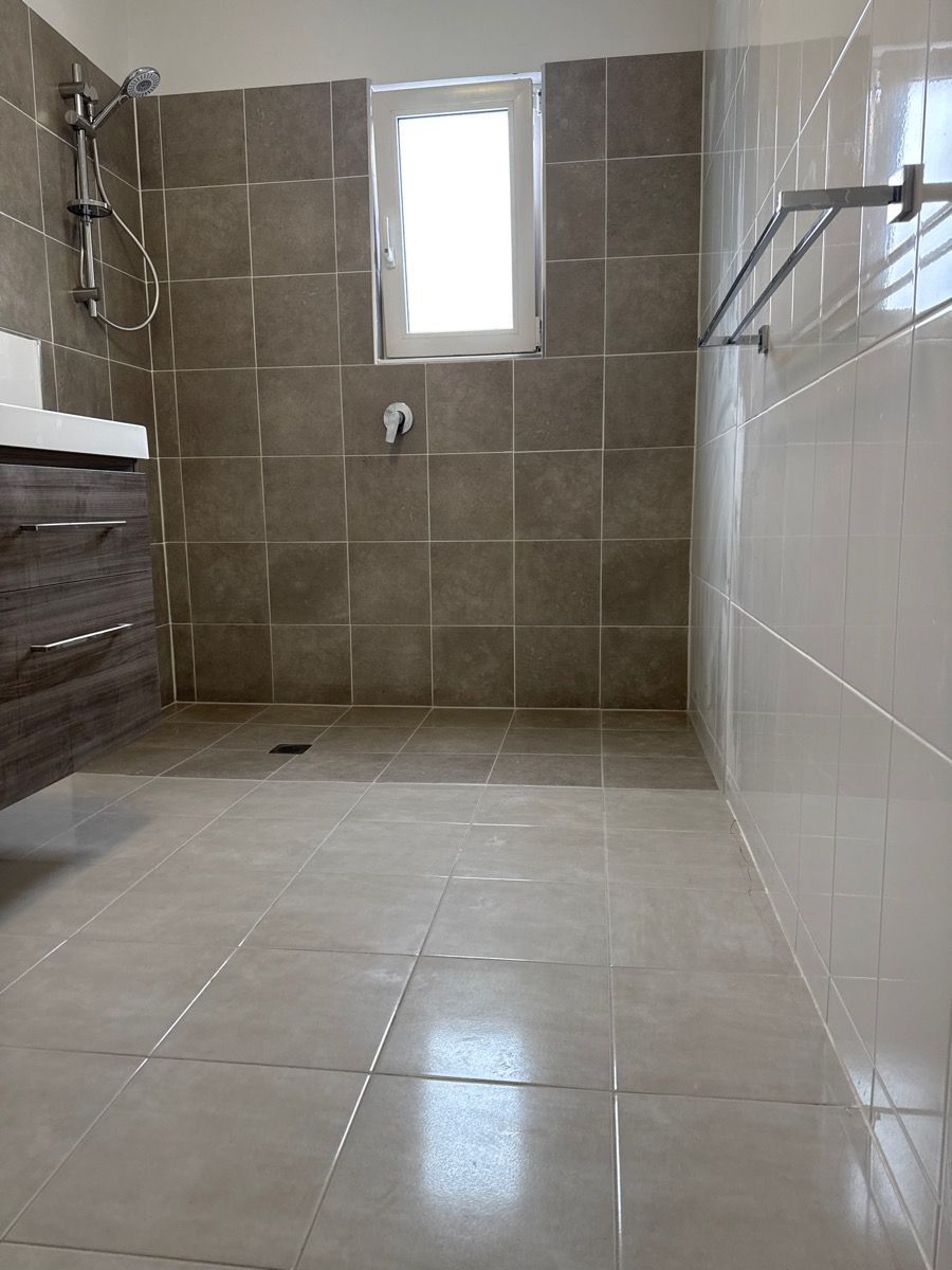 Empty tiled shower with beige walls, white floor tiles, grab bar, and small window.