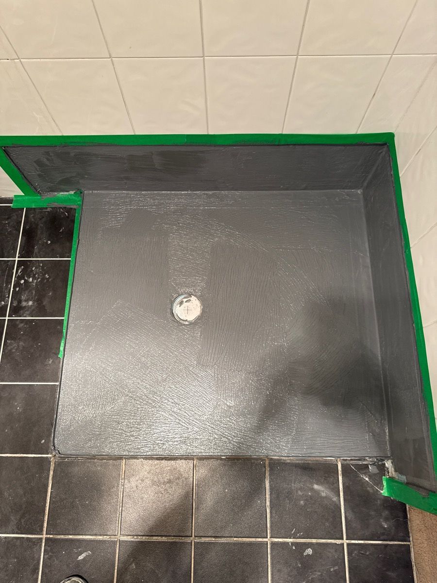 Black shower floor with a drain, bordered by green tape and surrounded by beige and black tiles.