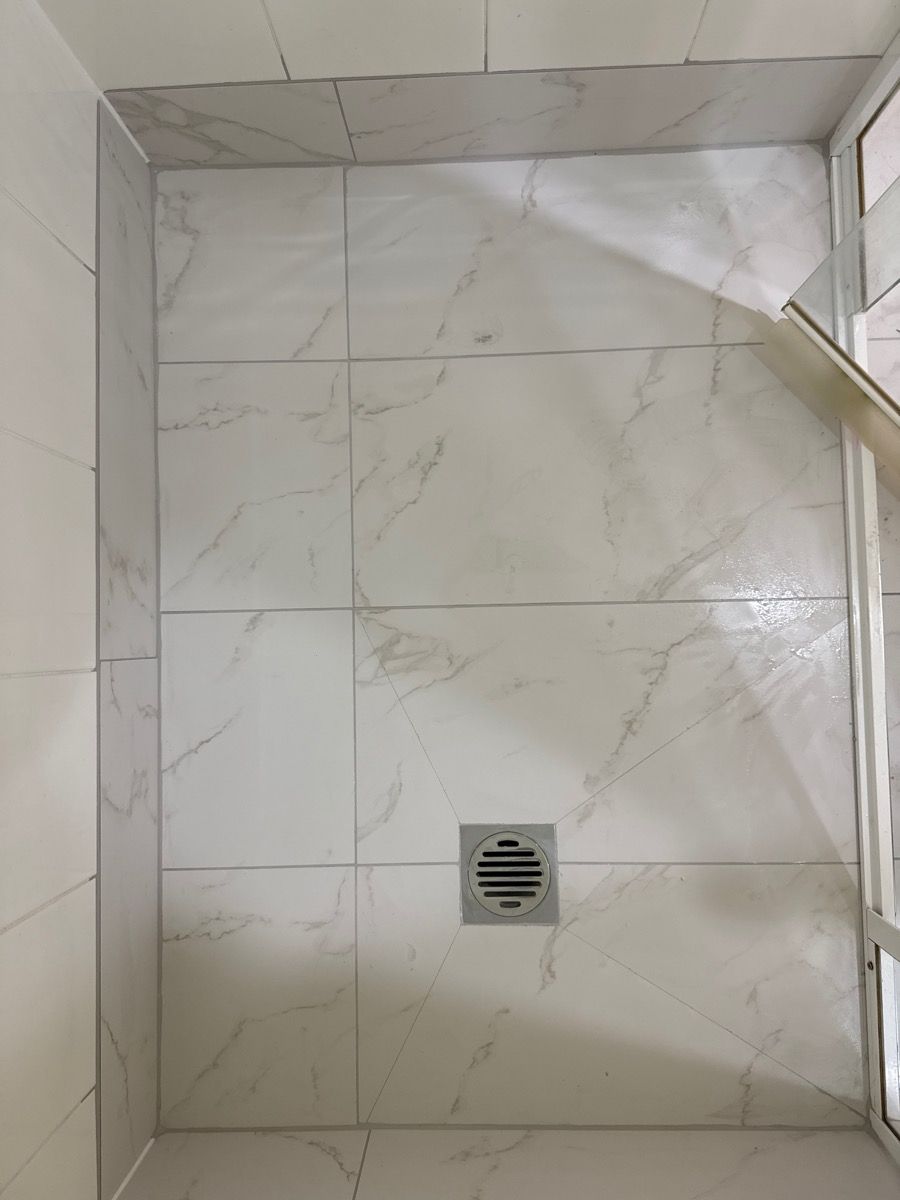 White tiled bathroom shower wall with a small square drain vent and glass door edge on the right