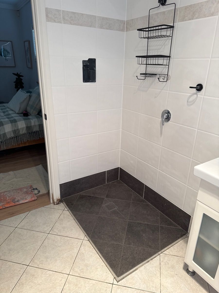 Corner shower area with white tile walls, dark floor tiles, and a wall-mounted shelf and shower fixtures.