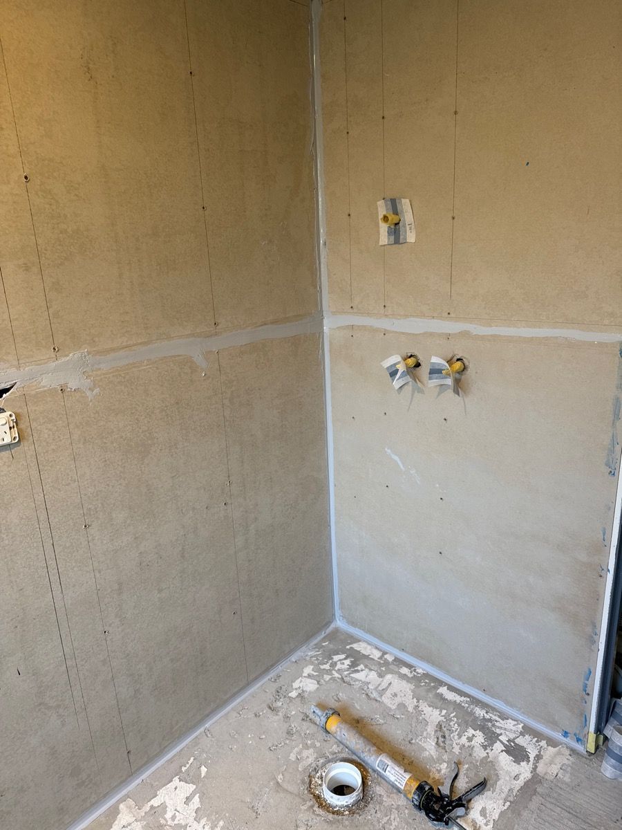 Unfinished bathroom corner with exposed drywall, plumbing hookups, and a toilet drain on the floor.