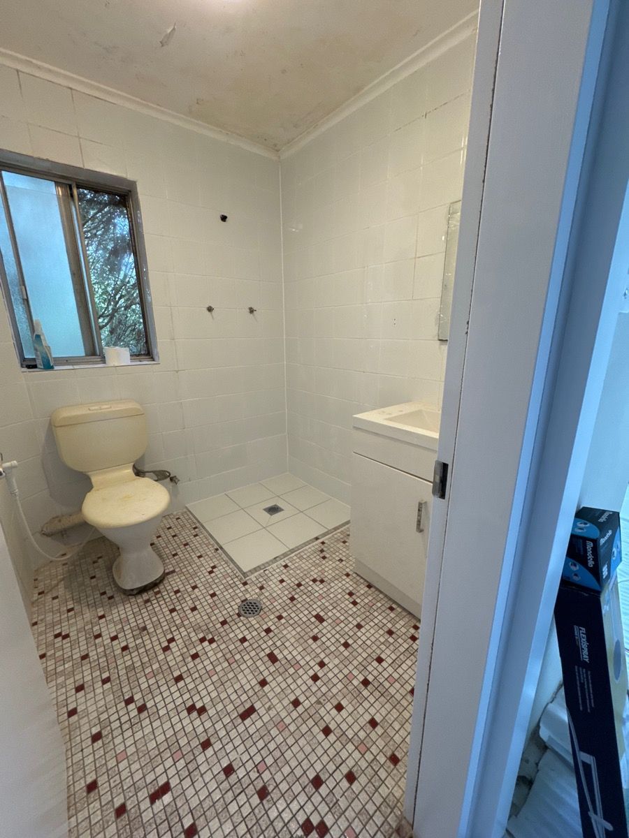 Small white bathroom with toilet, shower, sink cabinet, mosaic tile floor, and a window.