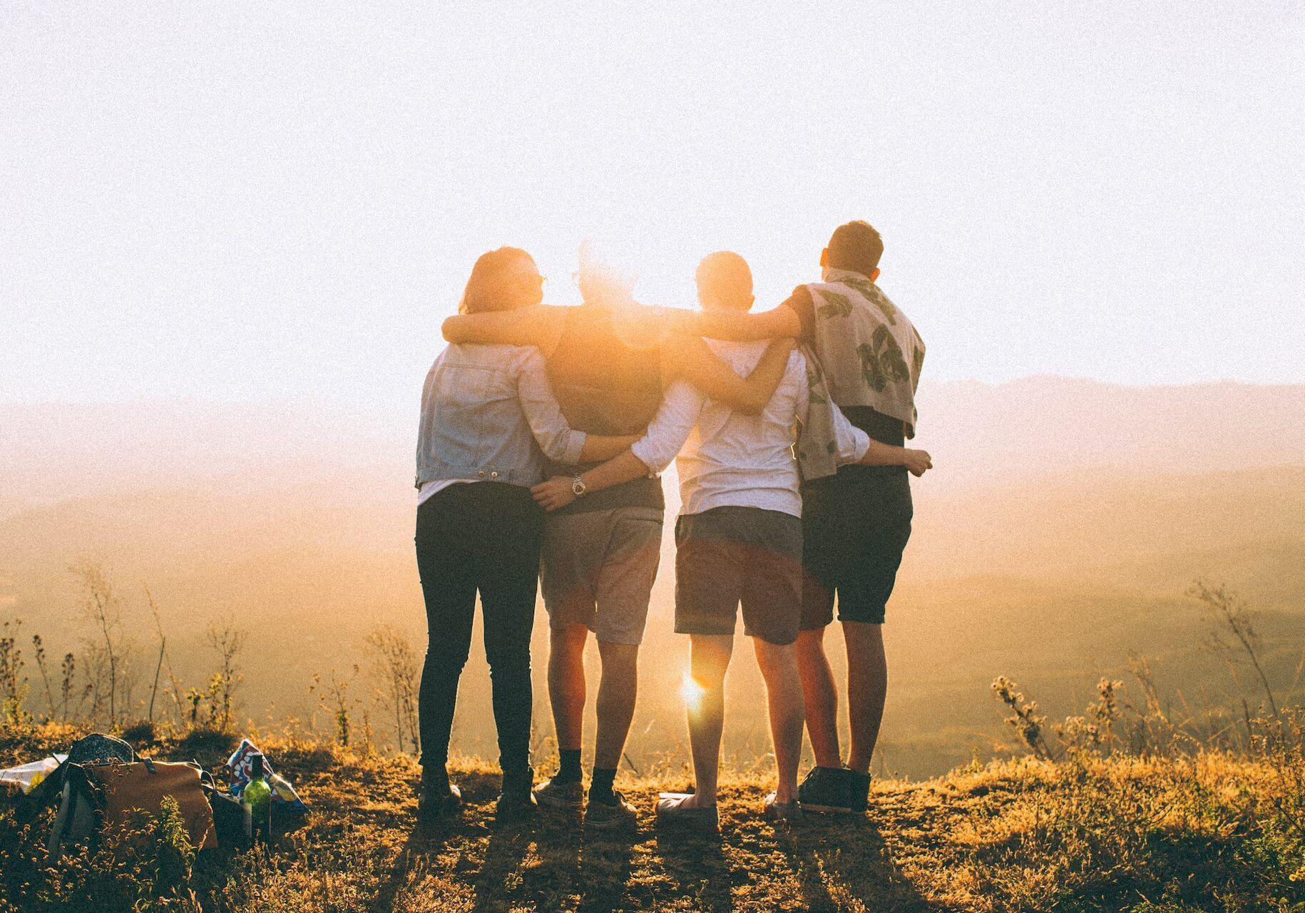 Four friends with arms around each other, silhouetted against a warm sunset over a mountain vista.