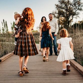 A family walks on a wooden bridge outdoors. A woman with red hair holds a baby. Others walk ahead, smil