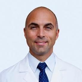 Smiling bald man in a white lab coat and blue tie against a light background