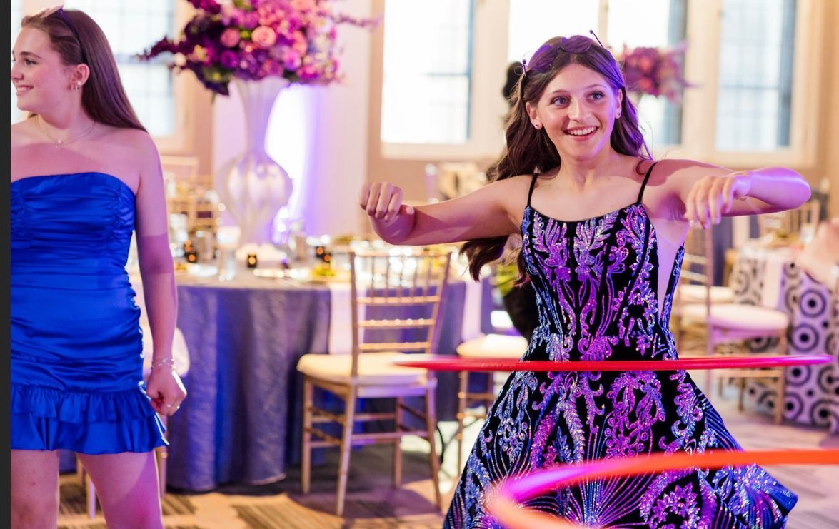 Two young women at a party; one in a black dress hula hoops, the other in a blue dress, purple floral arrangements.