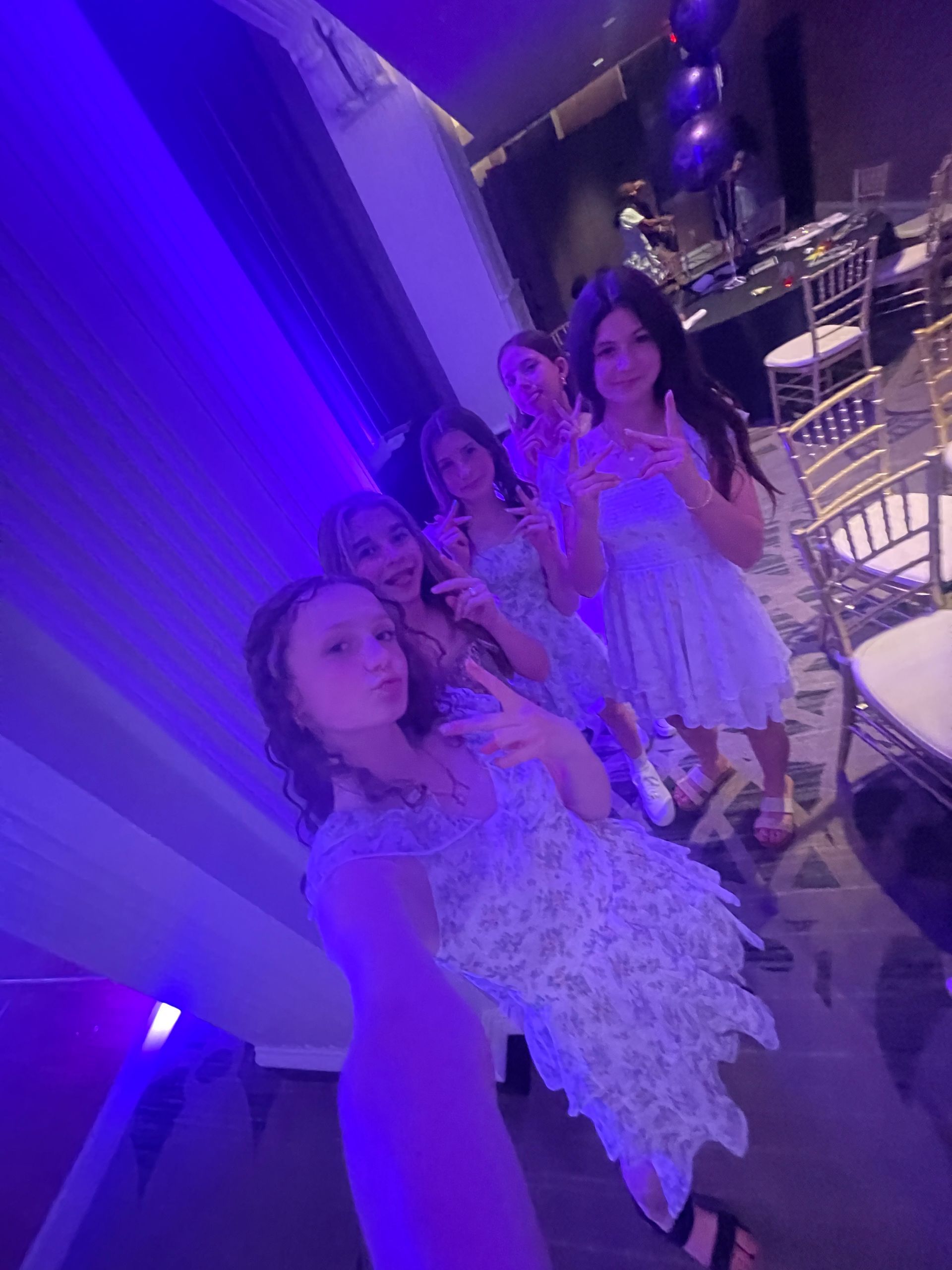 Five young women in light blue dresses pose for a selfie with peace signs, purple lighting.